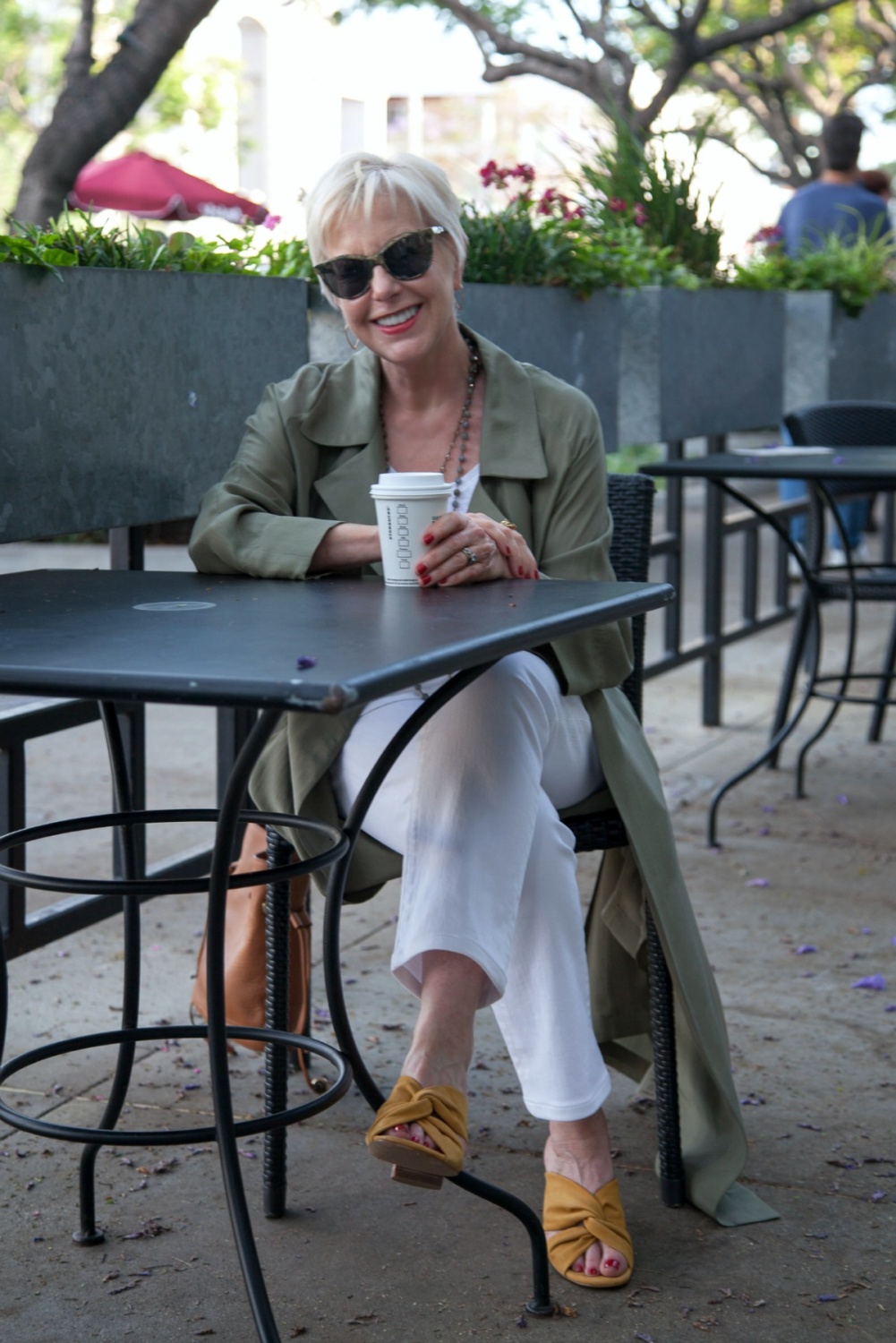 Pull up a chair! Style blogger Susan B. enjoys a coffee. Wearing olive silk trench over white jeans, yellow slide sandals. Details at une femme d'un certain age.