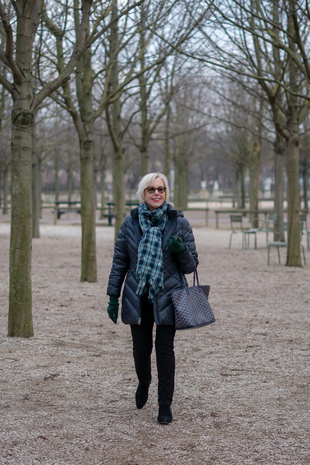 Travel outfit for Paris: style blogger Susan B. in Jardin du Luxembourg Paris, wearing a plaid scarf, puffer jacket, and Goyard tote. Details at une femme d'un certain age.
