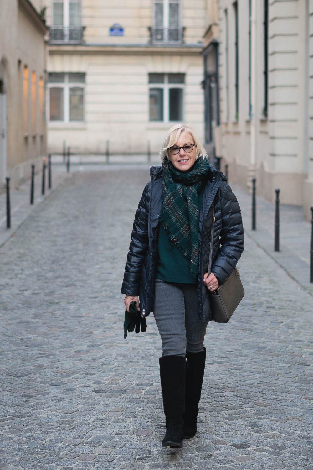 Winter travel outfit: Susan B. wears a navy puffer jacket, forest green top and scarf, grey jeans and black knee boots. Details at une femme d'un certain age.