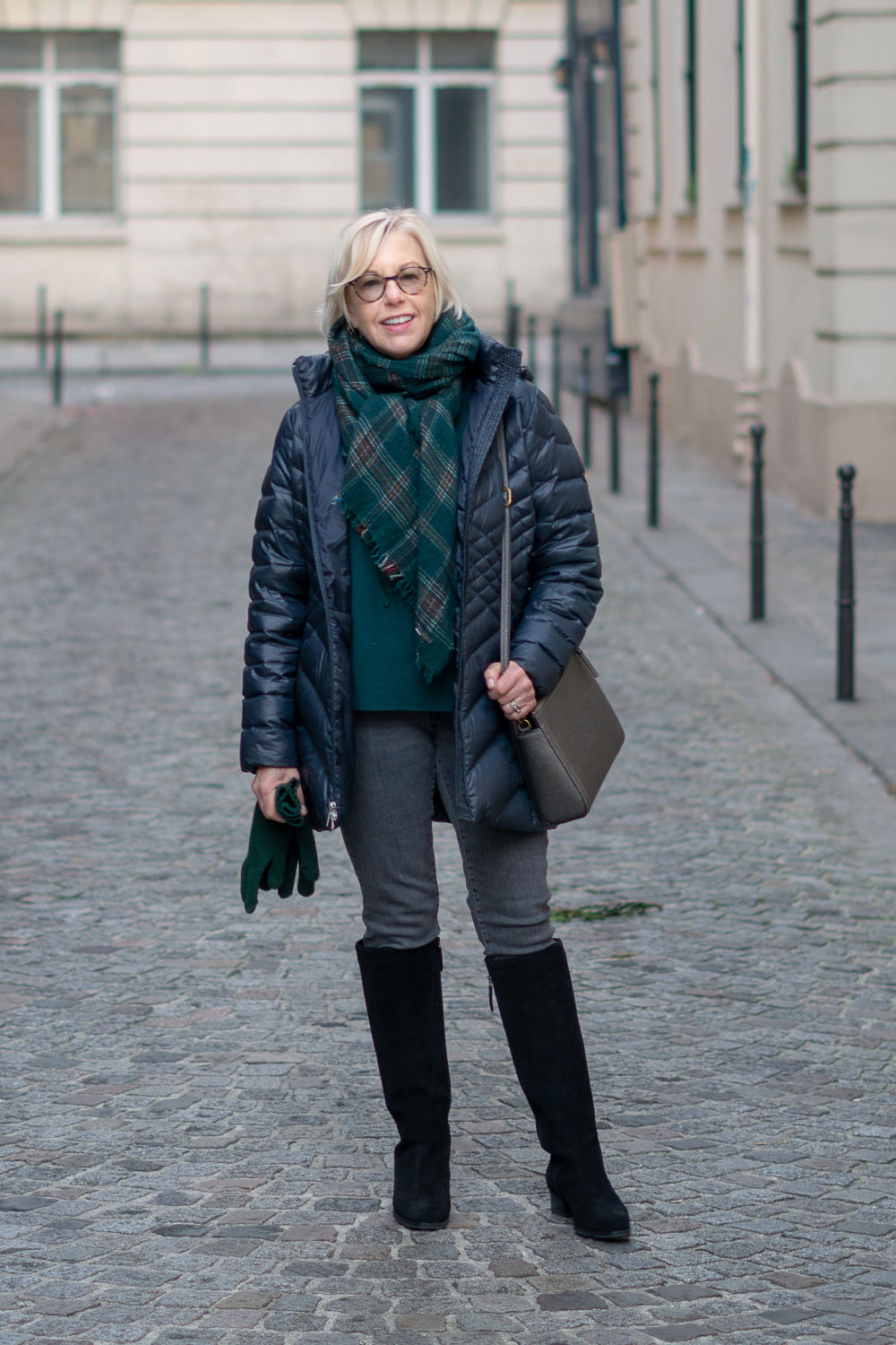 Winter travel outfit: Susan B. wears a navy puffer jacket, forest green top and scarf, grey jeans and black knee boots. Details at une femme d'un certain age.