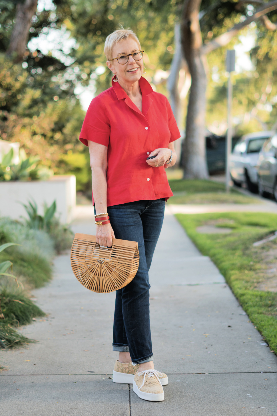 Susan B. of une femme d'un certain age wears a casual summer outfit with a red linen shirt, jeans, and raffia sneakers.