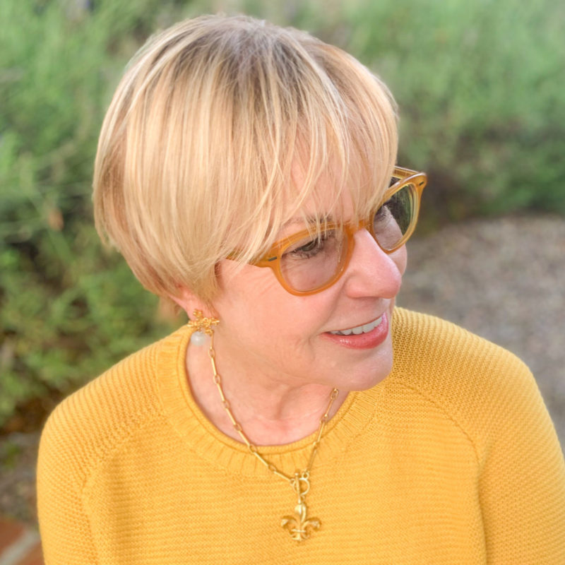 Detail: Susan B. wears French Kande gold earrings and fleur-de-lis medallion on a chain necklace. Info at une femme d'un certain age.