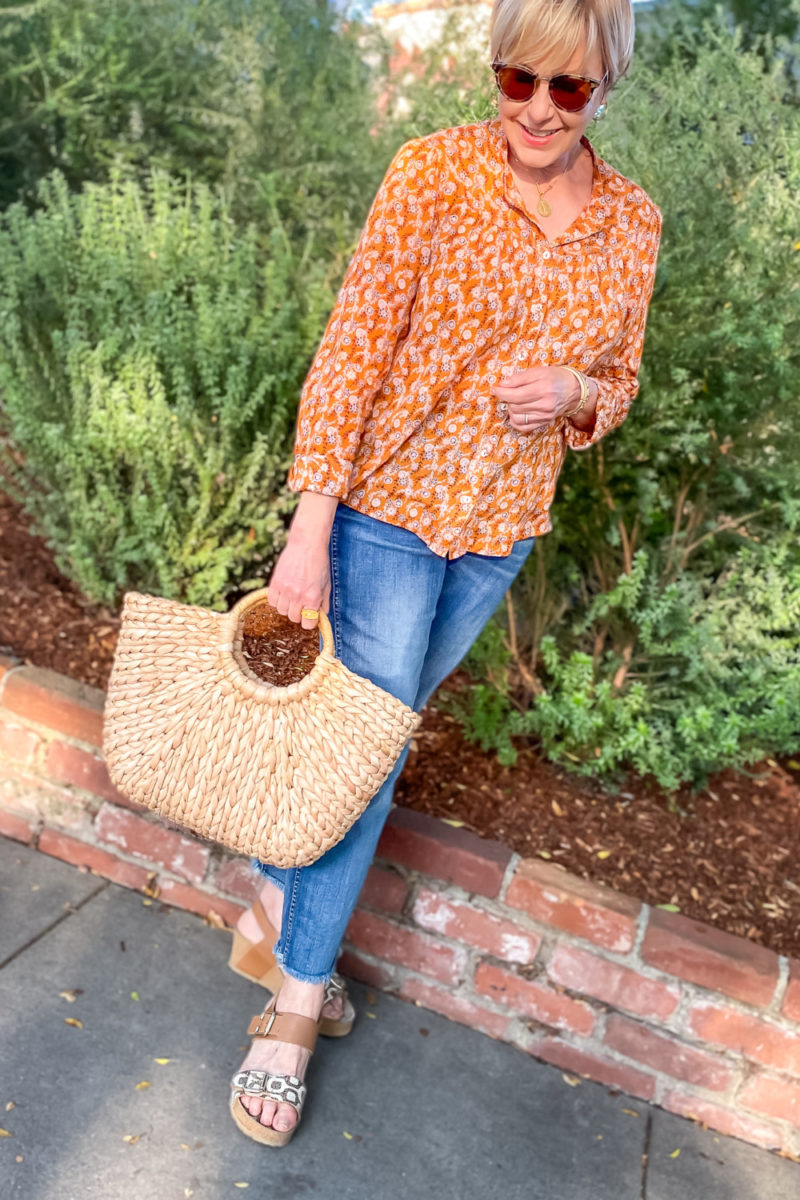 Susan B. wears a floral print top, cropped raw edge jeans, wedge sandals and a straw bag. Details at une femme d'un certain age.