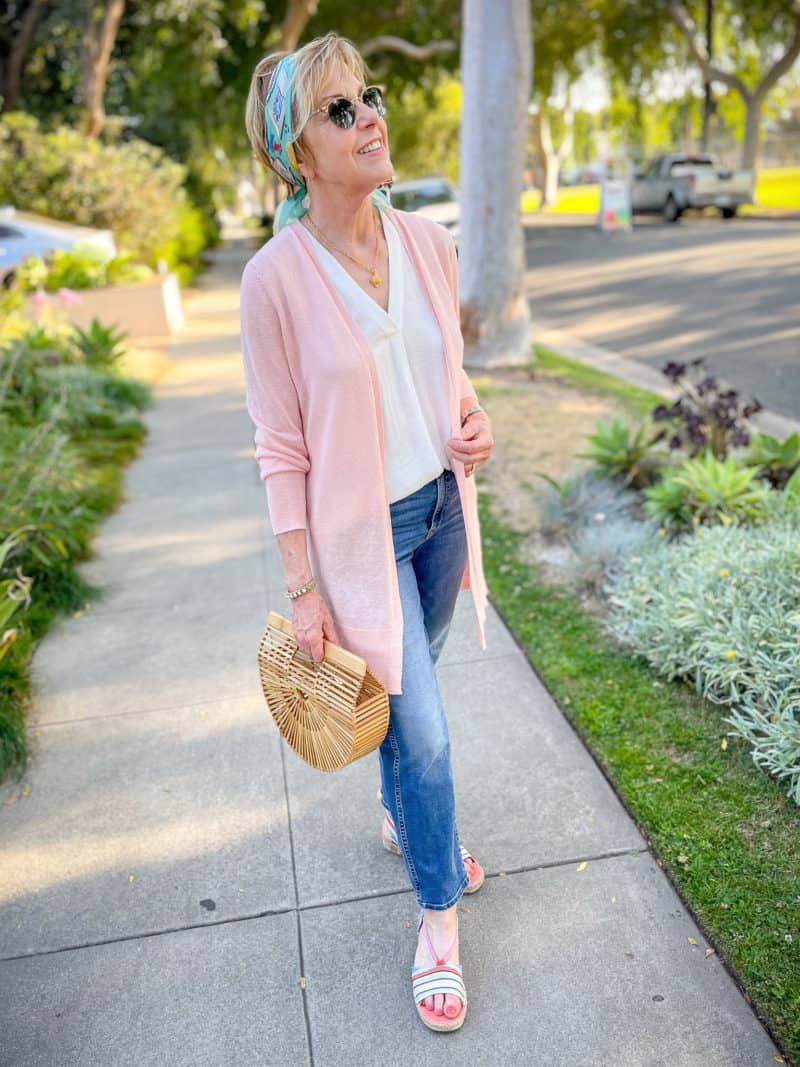A pink and blue summer outfit: Susan B. wears a pink linen cardigan, scarf headband, jeans, espadrille sandals, and carries a bamboo bag.