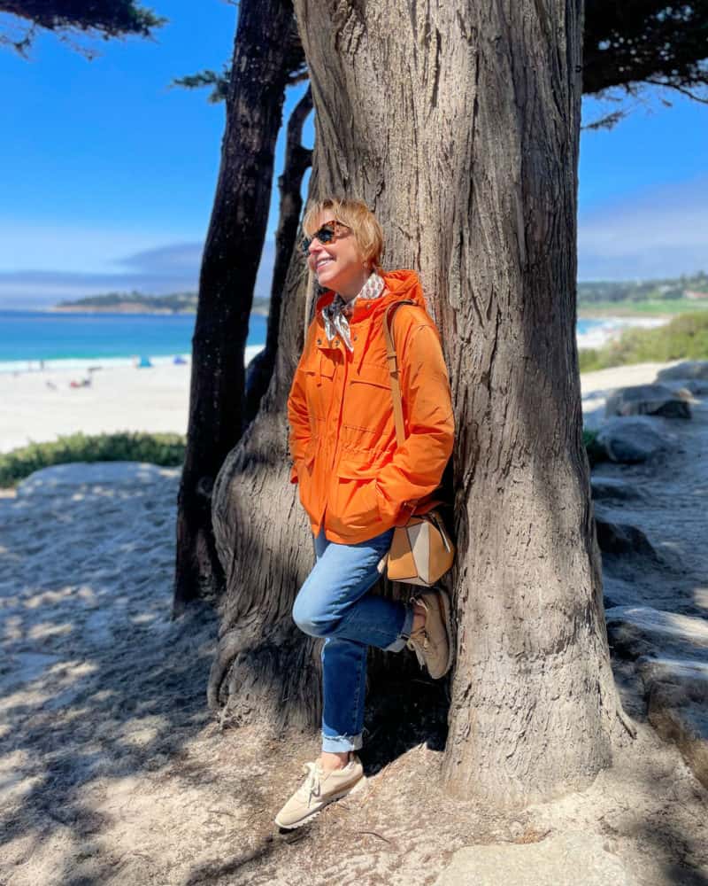 Susan leans on a tree at the beach in Carmel-by-the-Sea wearing an orange windbreaker and jeans.