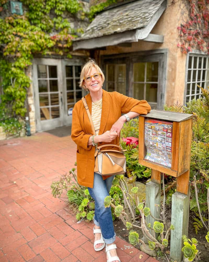 Susan B. wears a rust Madewell jacket and jeans, leaning against a flyer box in Carmel-by-the-Sea