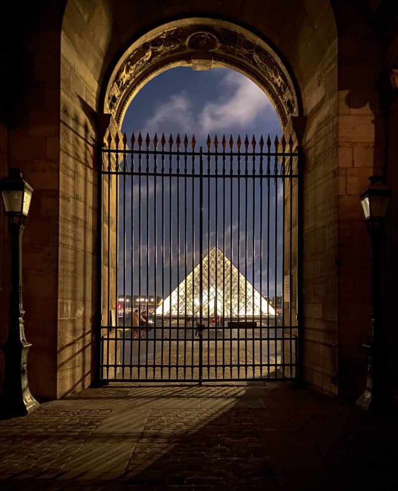 Pyramid in Louvre carousel lit at night, viewed through iron gate.
