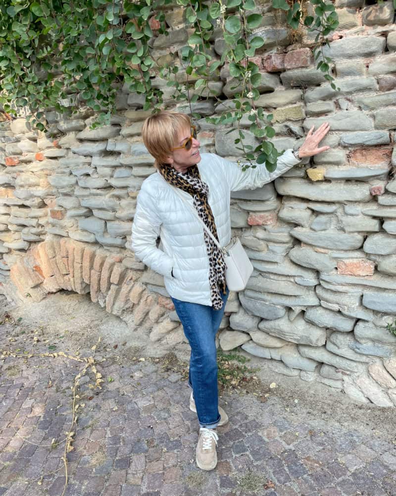 Italy travel outfit: Susan B. standing in front of a Medieval wall wearing an ivory down jacket, blue jeans, and a leopard print scarf.