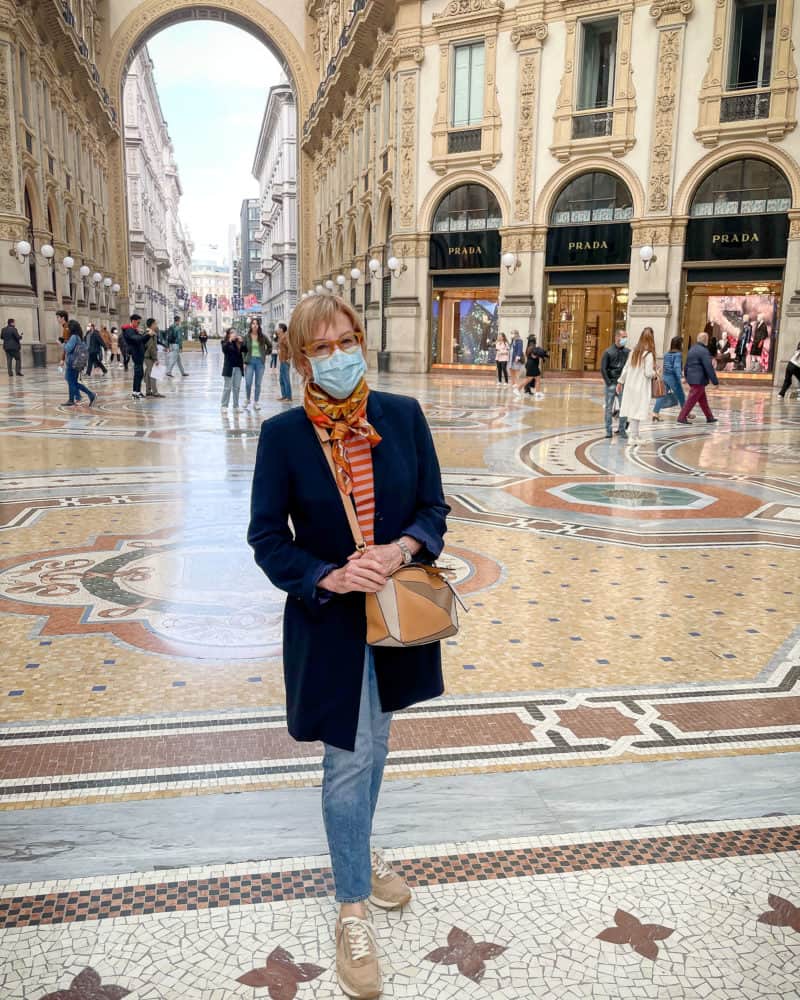 Susan B. in the Galleria in Milan wearing a long navy blazer, striped tee, silk scarf, jeans and sneakers.