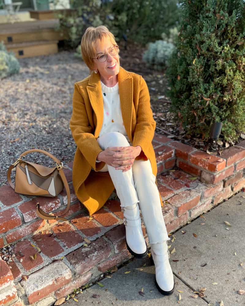 Susan B. sitting on steps wearing gold coat, knot detail sweater, ivory jeans, ivory lug-sole boots.