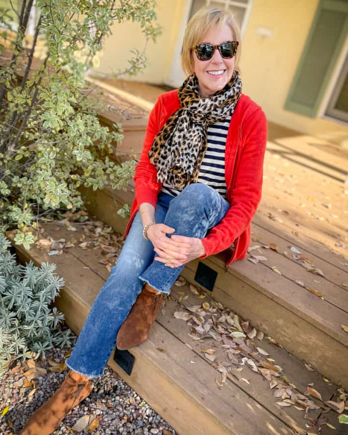 Susan B. sitting on wooden steps wearing leopard scarf, red cardigan, striped tee, jeans and boots.