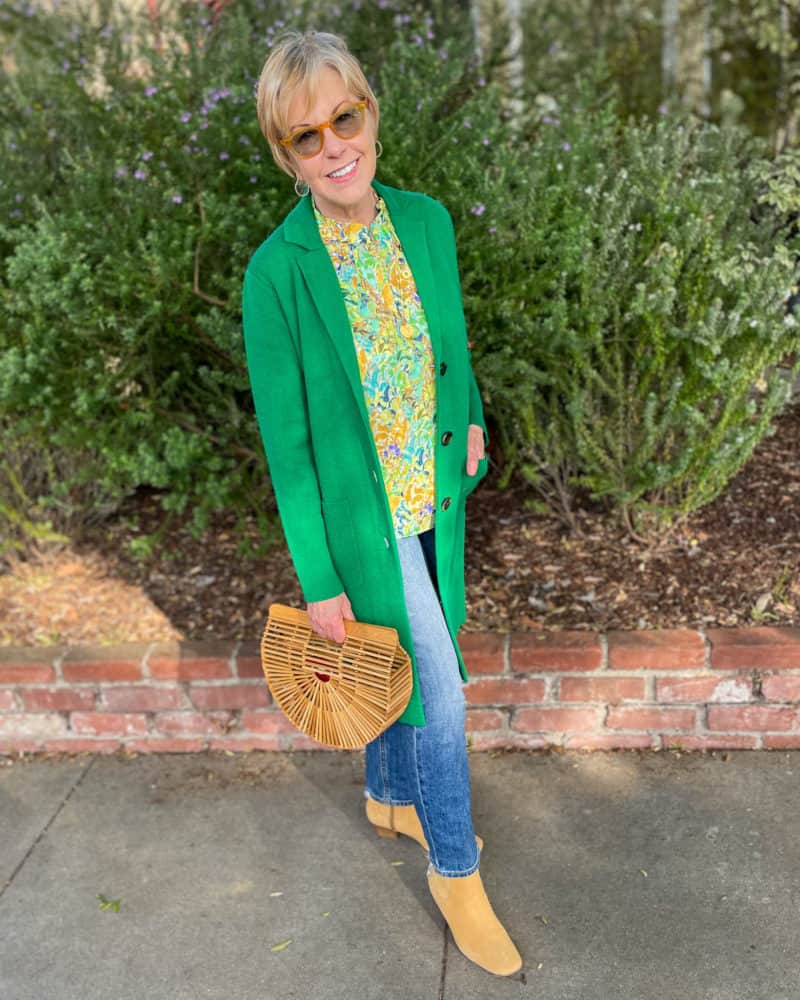 Susan B. wears a green coatigan, floral blouse, jeans, boots, and carries a bamboo bag.
