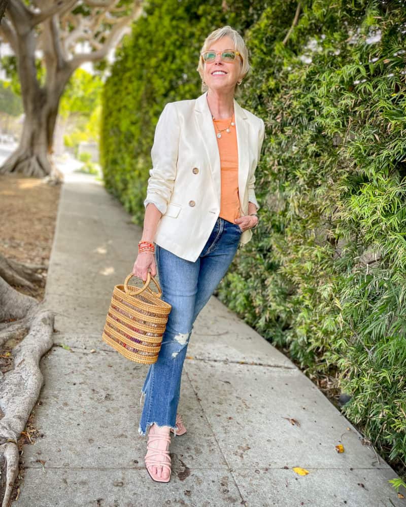 Susan B. wears a cream blazer, orange tee, casual pearl necklace, jeans, and carries a rattan bag.