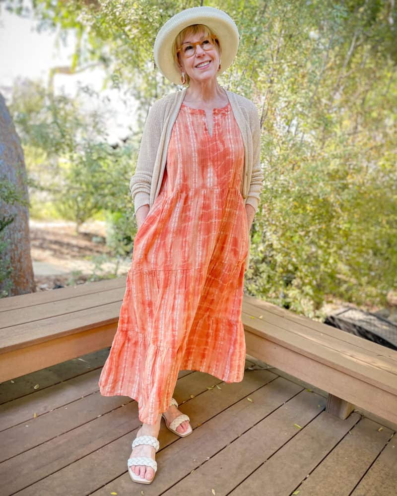 Susan B. wears a summer hat, tiered orange maxi dress, linen cardigan, and flat slide sandals.