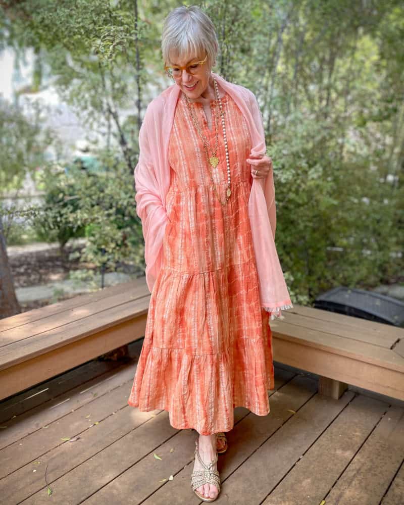 Susan B. wears an orange tiered maxi dress, sheer shawl wrap, gold and pearl layered necklaces, gold braided leather sandals.