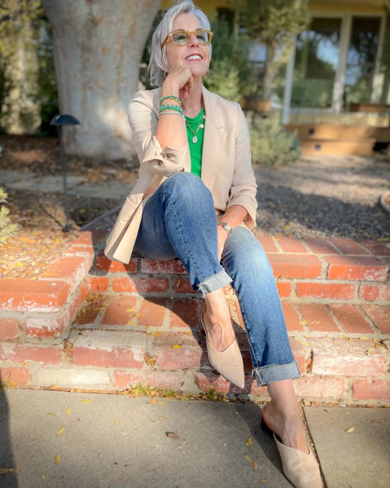 Susan B. sits on front steps wearing a light peach blazer, green tee, cuffed jeans and pointed toe mules.