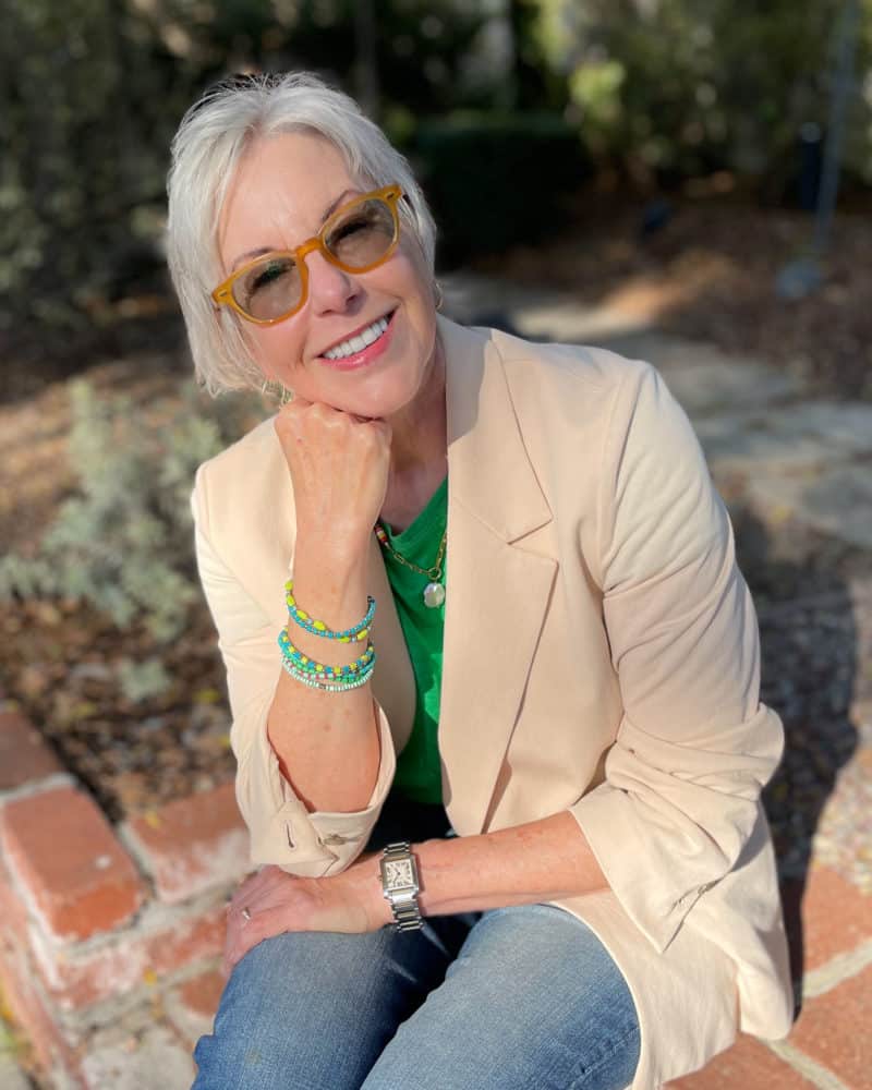 Susan B is seated, wearing a light peach jacket, green tee, green bracelets and blue jeans.