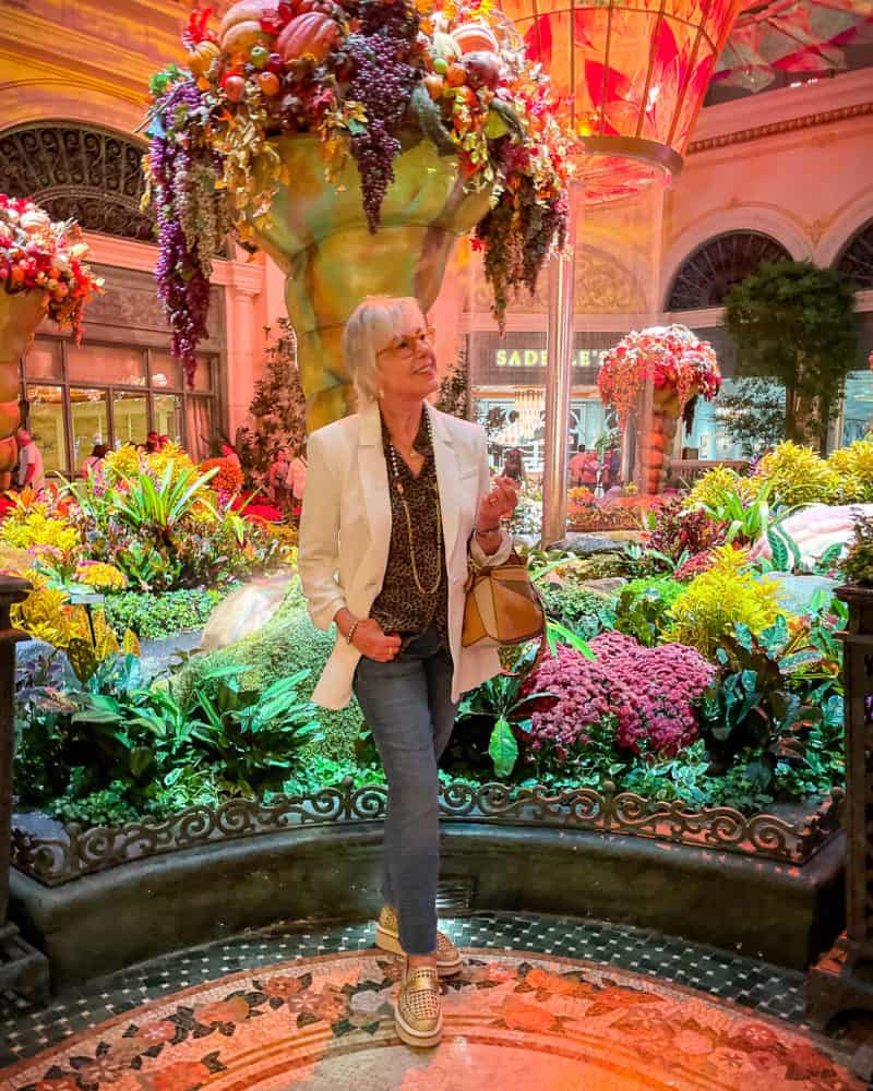 Susan B. stands in front of fall decorations at Bellagio Conservatory, wearing ivory jacket, leopard print top, jeans, gold loafers.