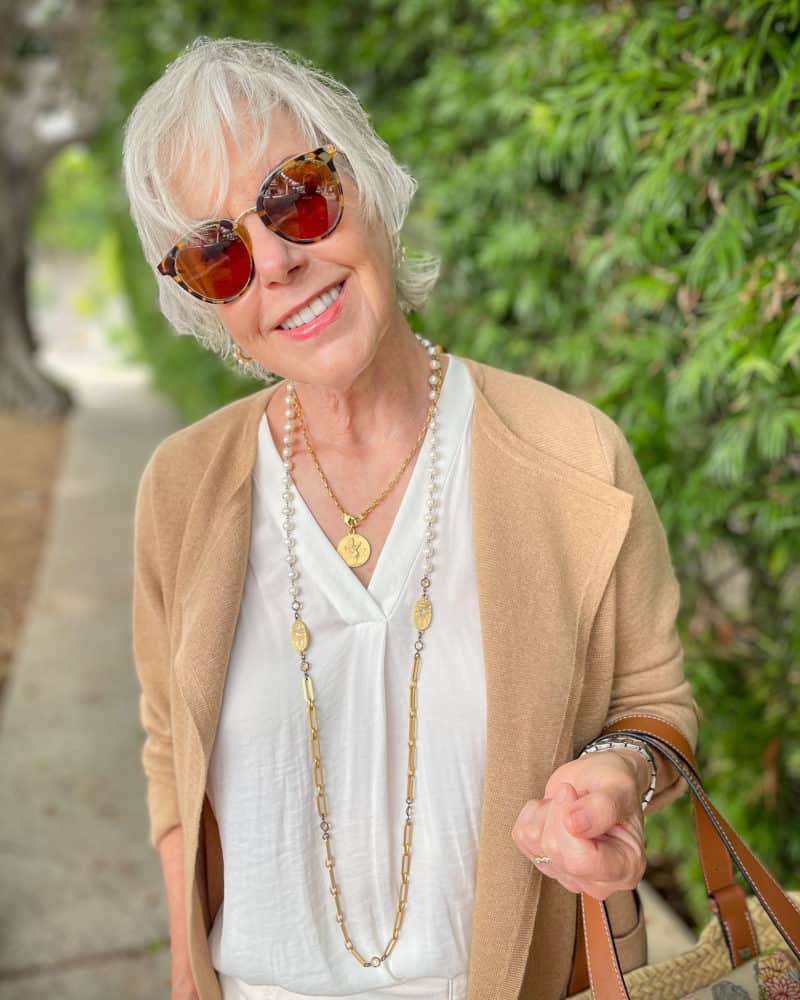 Detail: Susan B. wears gold & pearl French Kande necklaces, a v-neck top, sunglasses & camel sweater jacket.