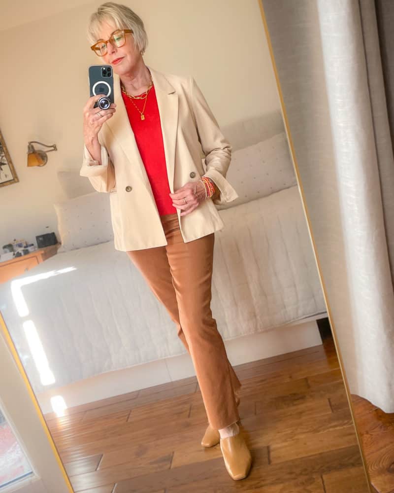 Susan B. wears a light peach jacket, red sweater, tan coated denim jeans, camel mules, gold necklaces.