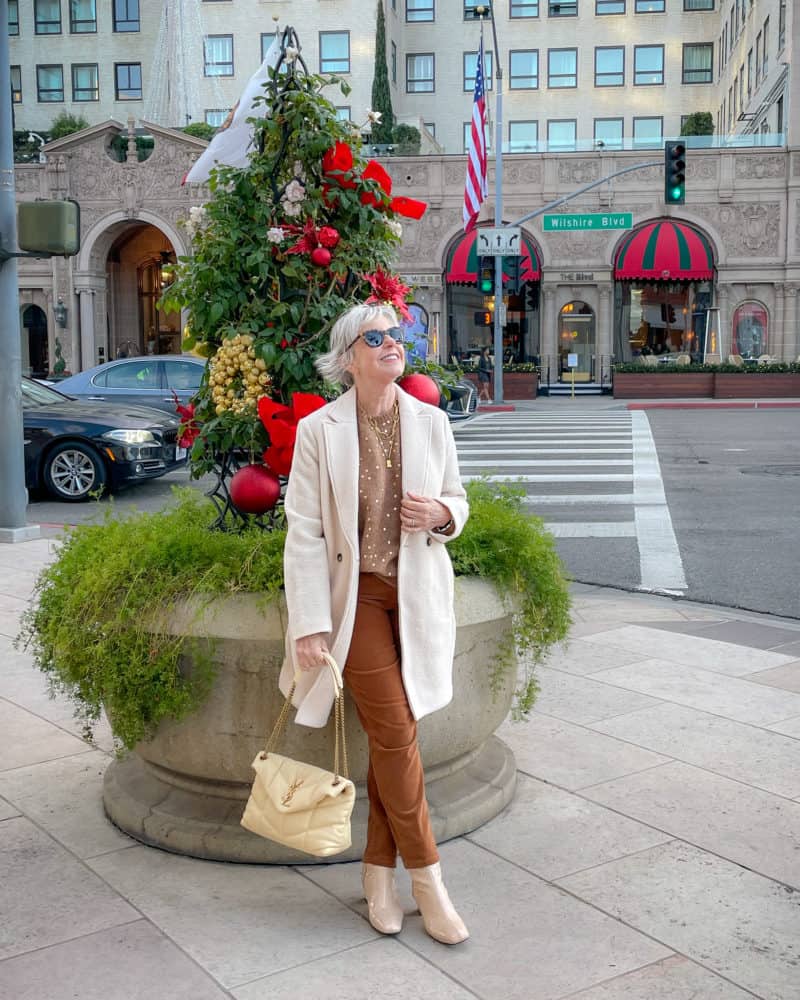 Susan B. in front of a holiday planter in Beverly Hills. Wears an ivory topcoat, brown metallic dot sweater, brown corduroy pants, light yellow Saint Laurent Loulou puffer bag.