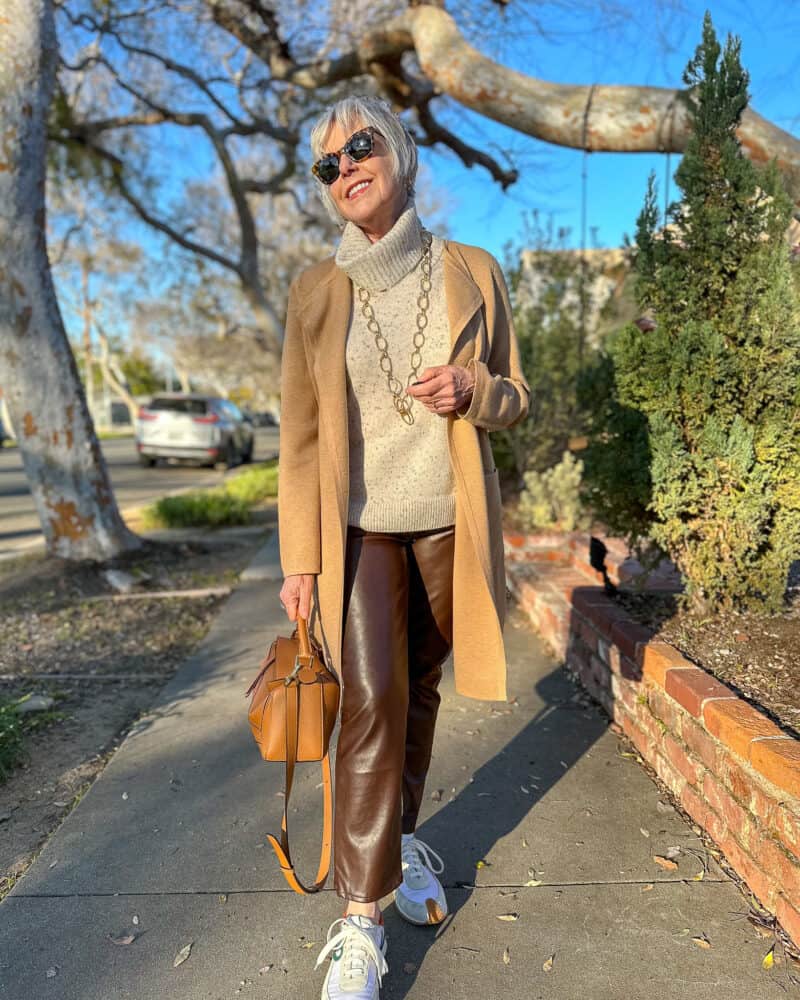 Susan B. wears a casual faux leather pants outfit with a sweater vest, long sweater jacket and sneakers.