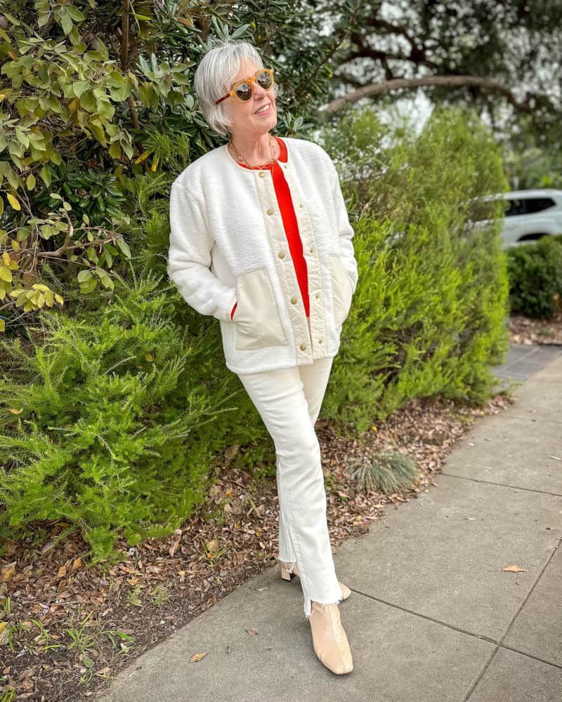Susan B. wears a winter white outfit with an sherpa fleece jacket, orange sweater, off-white jeans, patent leather boots.