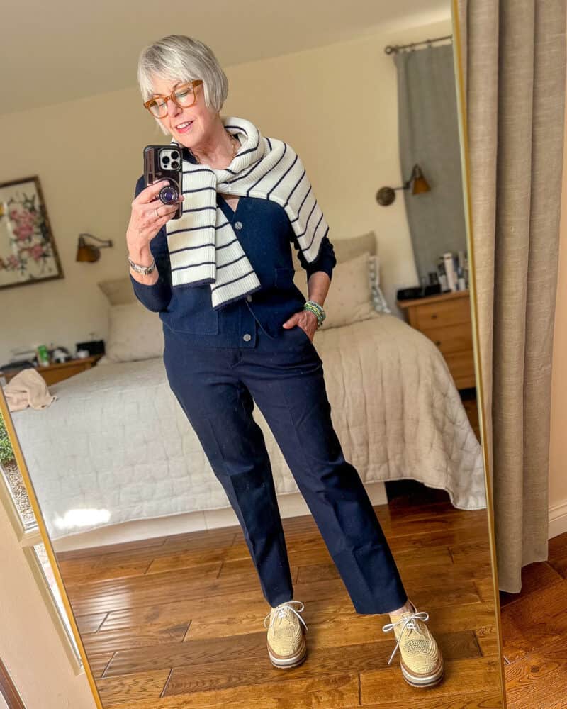 Susan B. wears a navy sweater and trousers, with a striped sweater ties over the shoulders, and raffia oxfords.