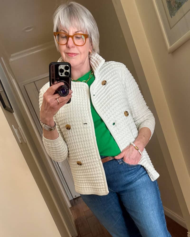 Susan B. wears an ivory textured sweater jacket, green cashmere tee, brown belt, pearl bracelet, blue jeans.