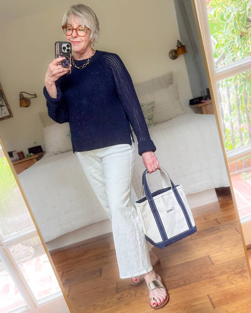 Susan B. wears a textured navy cotton sweater, white wide leg jeans, gold sandals, carries a canvas tote.