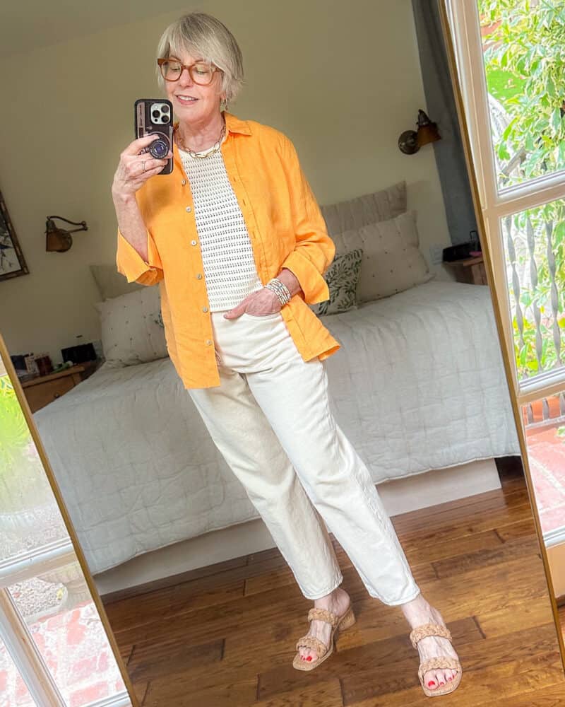 Susan B. wears white boyfriend jeans with a crochet vest, orange linen shirt, cork effect sandals.