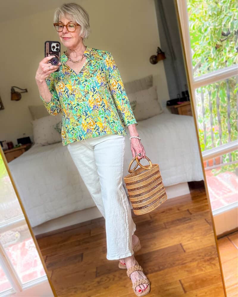 Susan B. wears a floral print silk blouse, wide leg white jeans, cork sandals, carries a rattan bag.