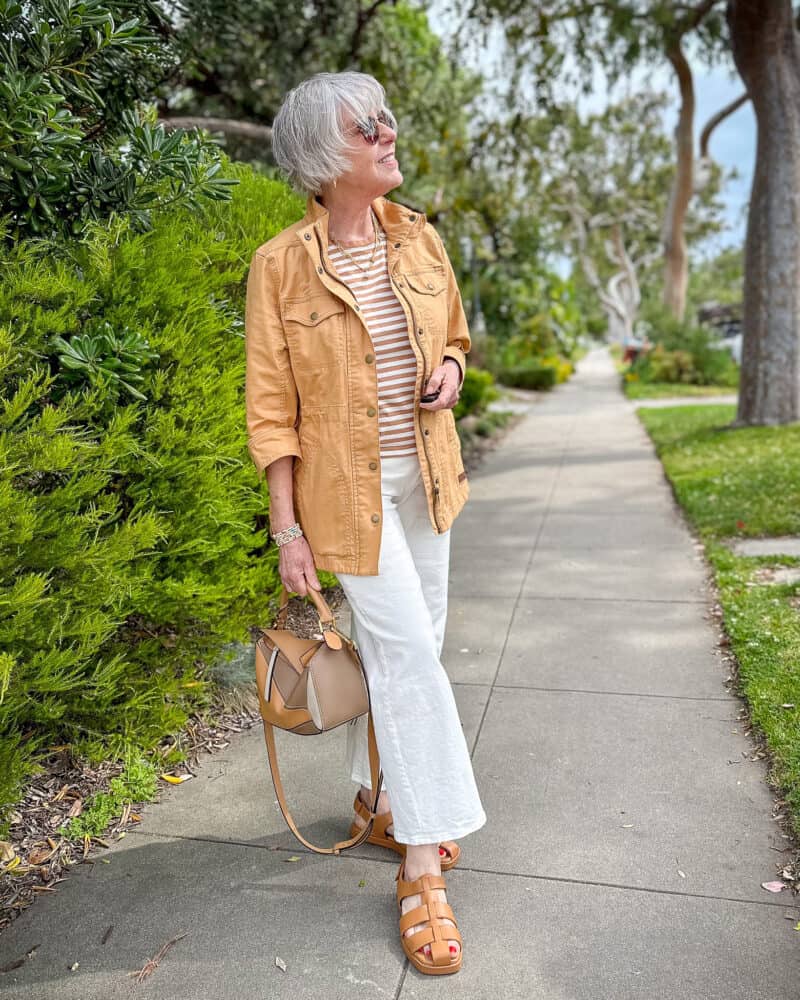 Susan B. wears a casual caramel and white outfit with a utility jacket, striped tee, fisherman sandals.