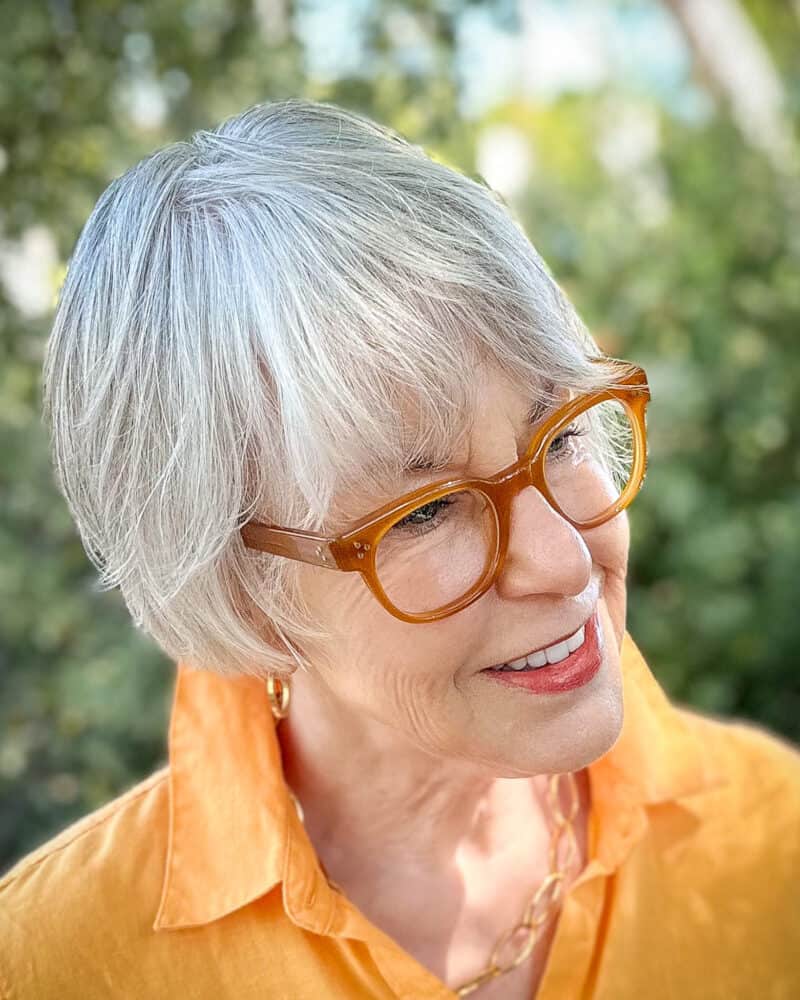 Susan B. with gray hair cut in a layered bob, wears an orange shirt and gold jewelry.