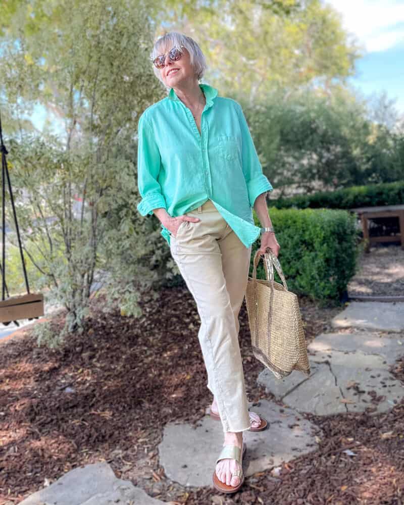 Susan B. stands in a garden wearing an aqua shirt, tan chinos, gold sandals, carrying a raffia tote