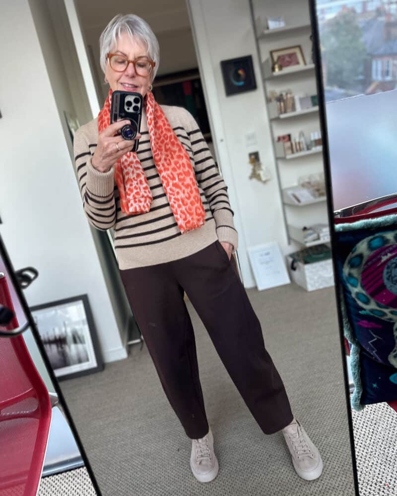 Susan B. takes a mirror selfie wearing a coral leopard print scarf, striped sweater, brown knit pants and sneakers.