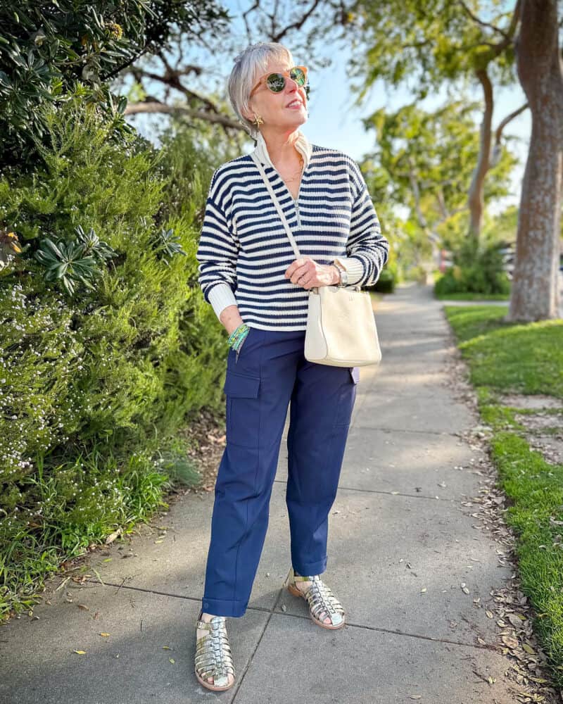 Susan B. wears a zip-neck striped sweater, navy cargo pants, metallic fisherman sandals, and an ivory crossbody bag.