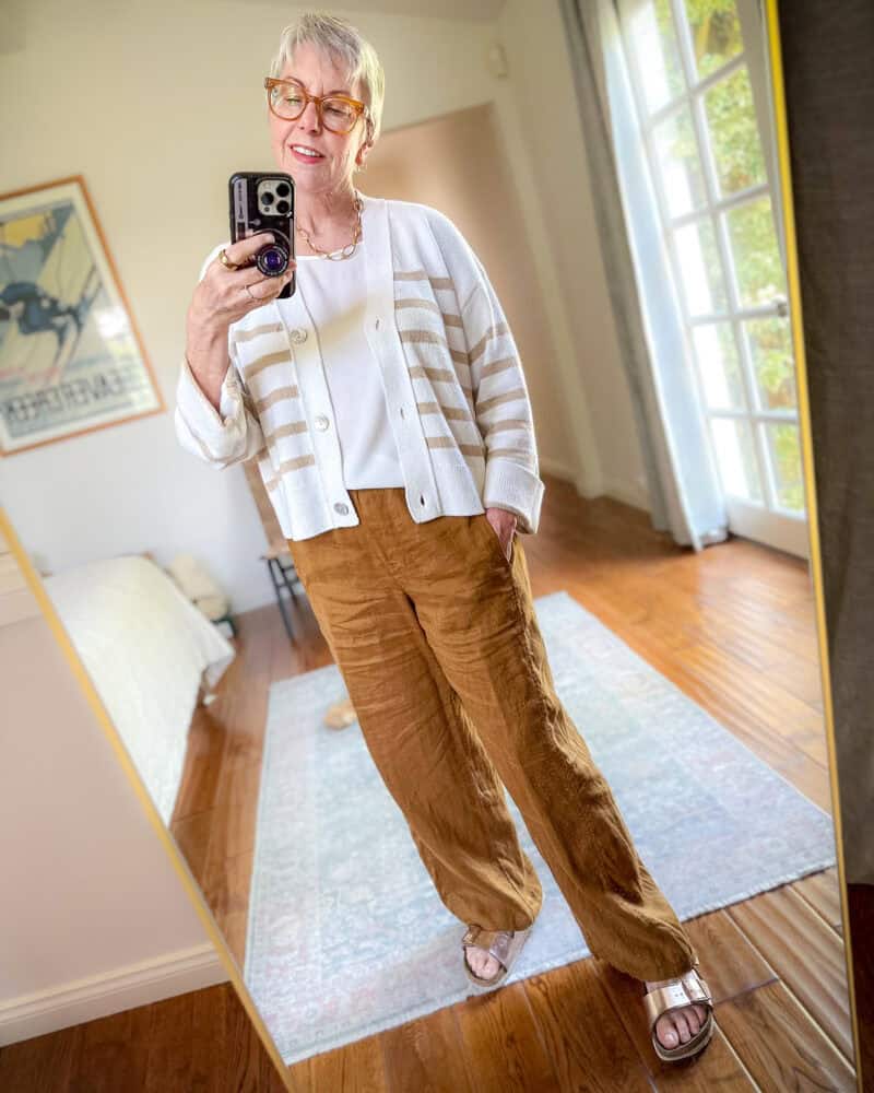 Susan B. wears a white and beige striped cardigan, off-white top, brown linen pants and gold Birkenstock sandals.
