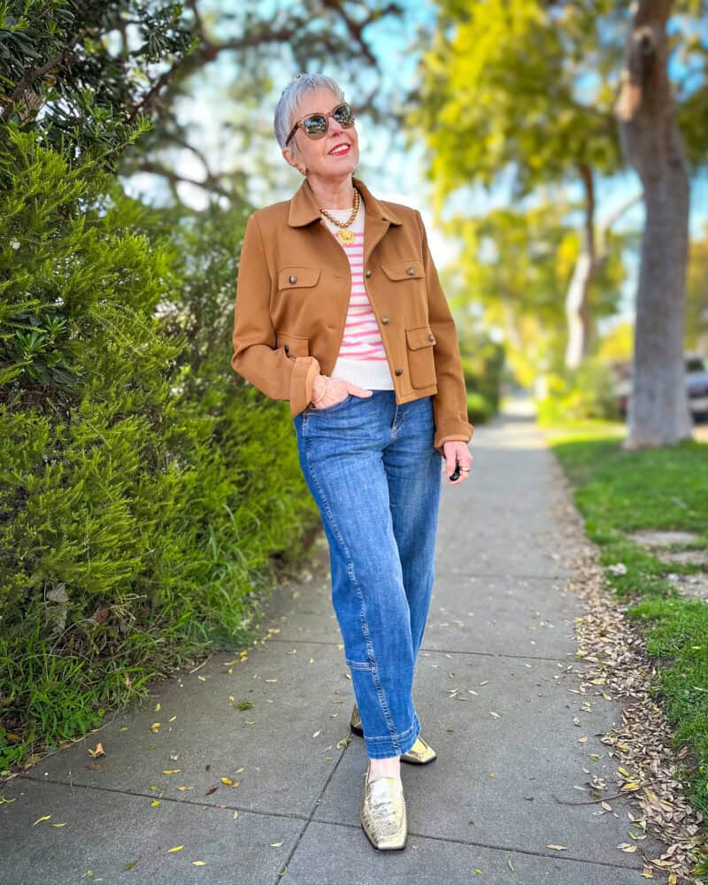 Susan B. wears a cropped brown utility jacket, pink stripe cashmere tee, gold pearl necklace with medallion, barrel leg jeans, and gold loafers.