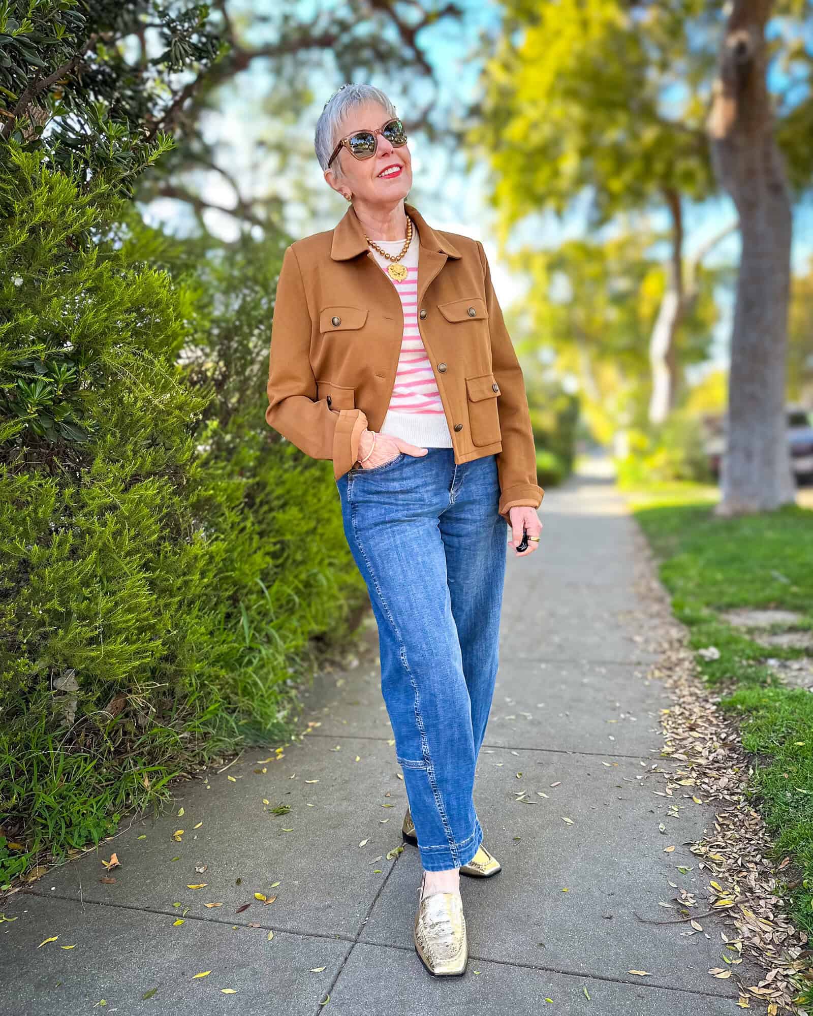 Susan B. wears a cropped brown utility jacket, pink stripe cashmere tee, gold pearl necklace with medallion, barrel leg jeans, and gold loafers.