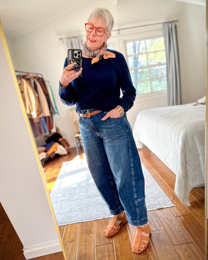 Susan B. wears a navy sweater with medium wash barrel leg jeans, a cotton bandana, and fisherman sandals.