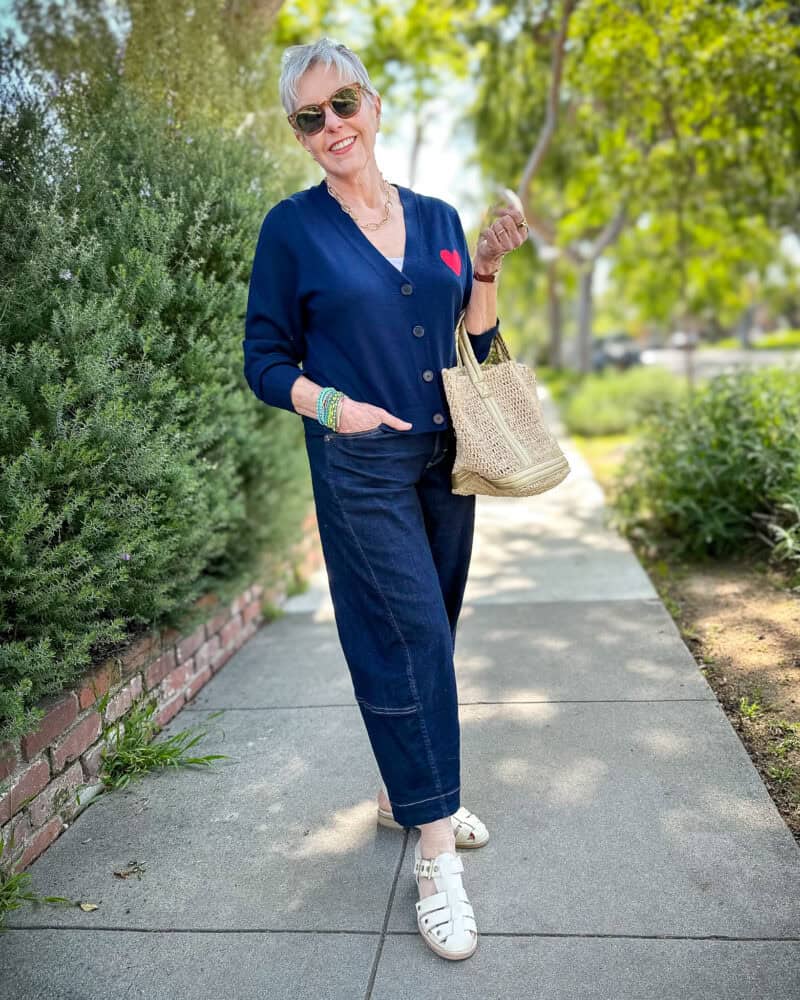 Susan B. wears a navy cardigan with red heart detail, dark wash barrel jeans and ivory fisherman sandals, carries a raffia tote bag.