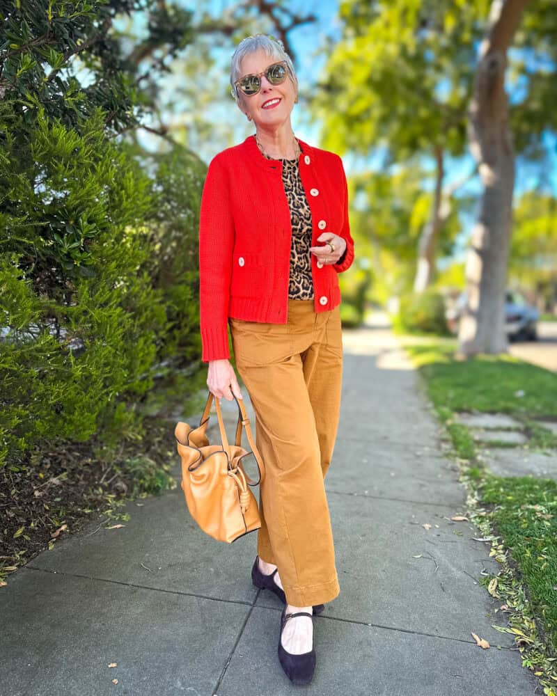 Susan B. wears a red cotton cardigan, leopard print cashmere shell, tan pants, dark brown Mary-Jane flats, carries a tan leather Loewe Flamenco bag.