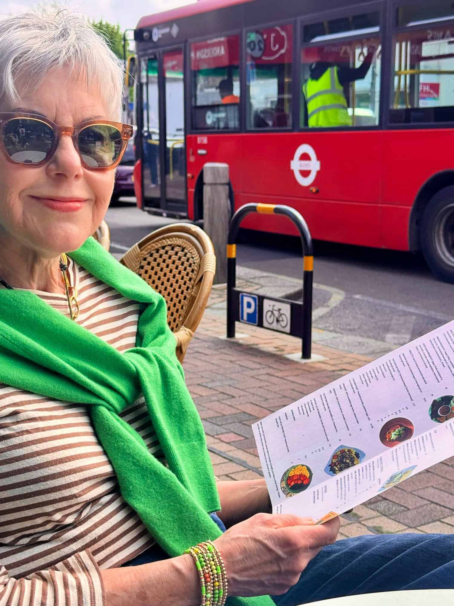 Susan B. reads the menu at an outdoor cafe with a red bus in the background.