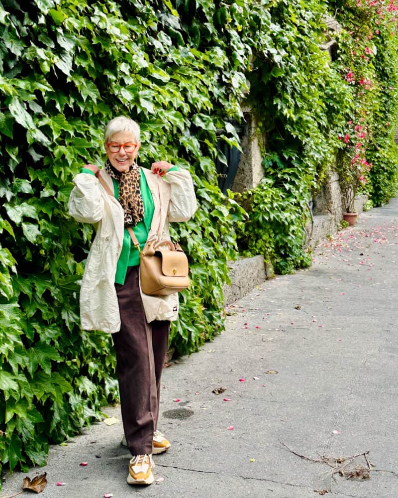 Susan B. wears a Weekend Max Mara packable rain jacket, green sweater, leopard print scarf, brown pants, color-block sneakers.