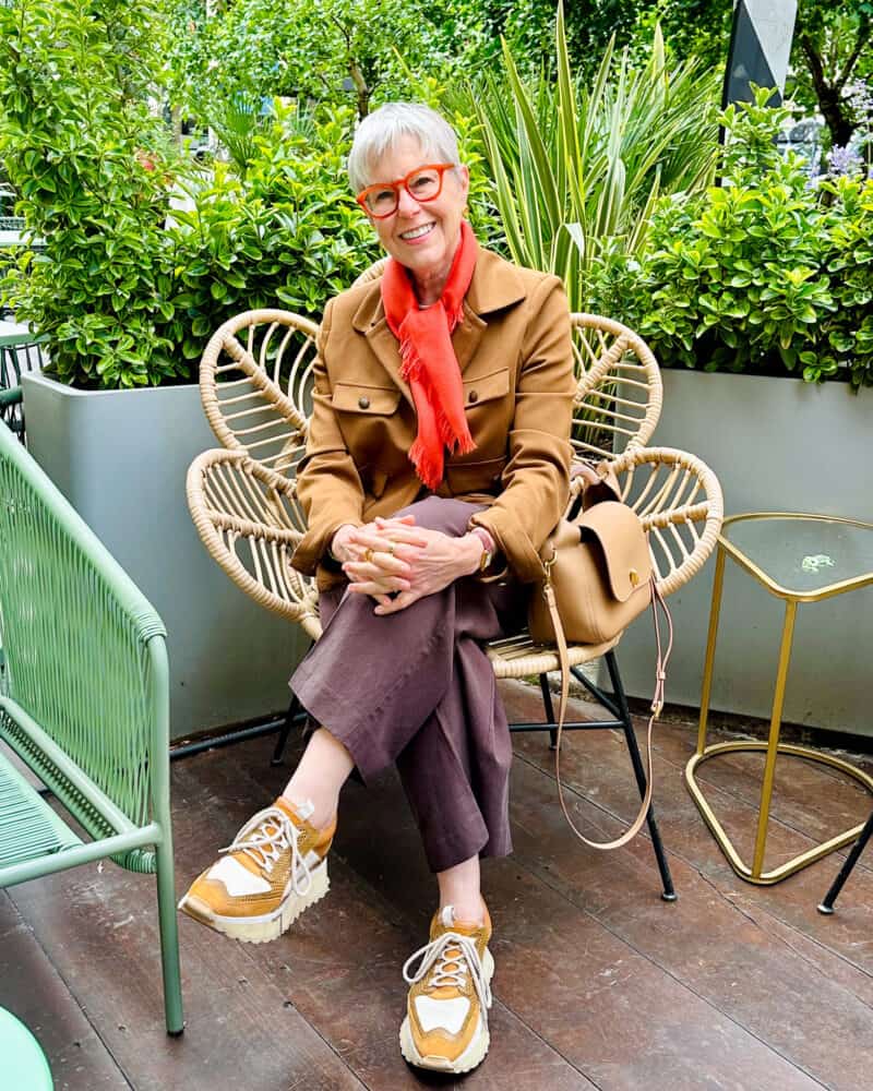 Susan B. sits in a decorative rattan chair, wearing an orange scarf, brown jacket, brown pants, platform sneakers.