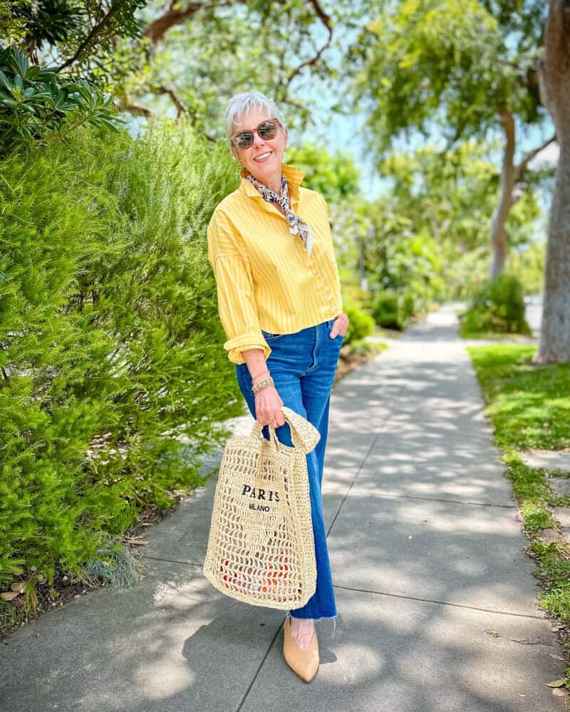 Susan B wears a cropped yellow shirt, patterned bandana, jeans, pointed to flats, carries a straw tote with "Paris/Milano" writing.
