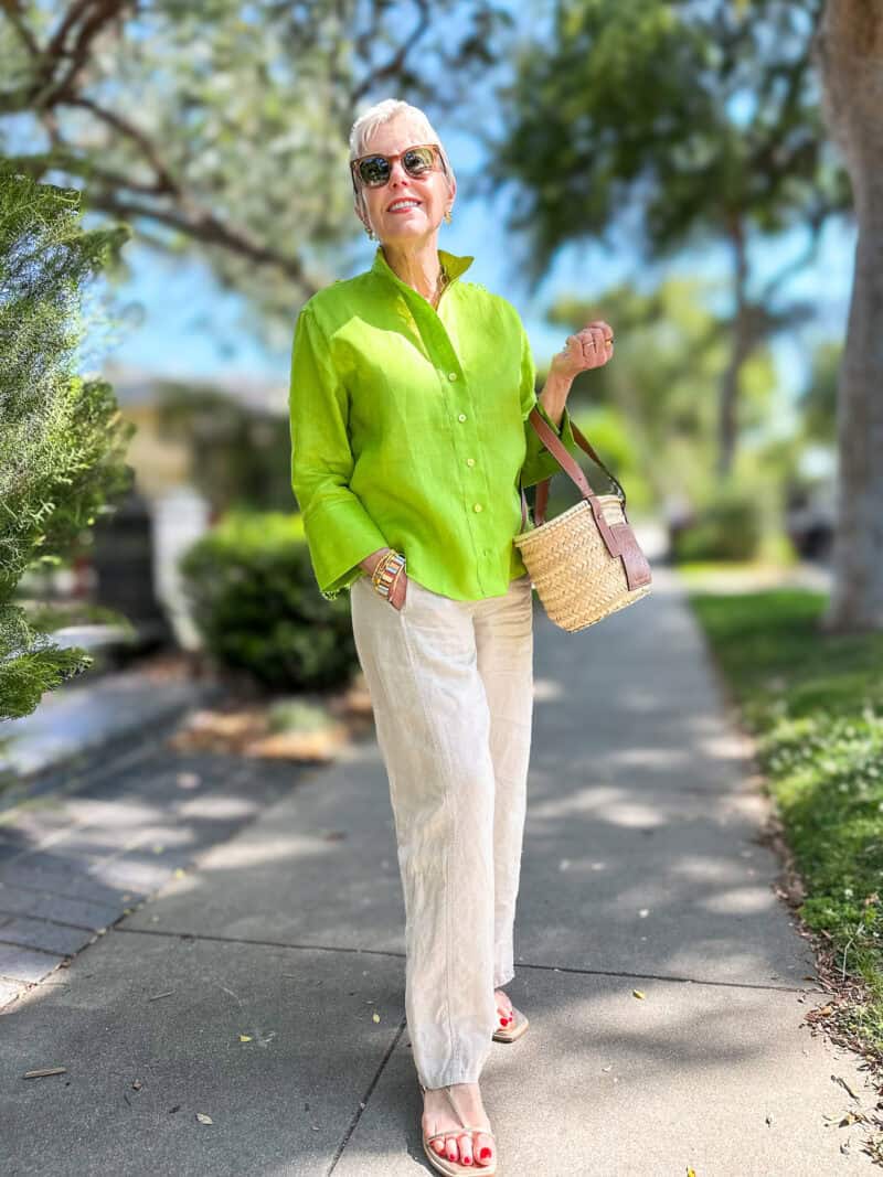Susan B. wears a green linen shirt, undyed linen trousers, gold sandals, carries a basket bag.