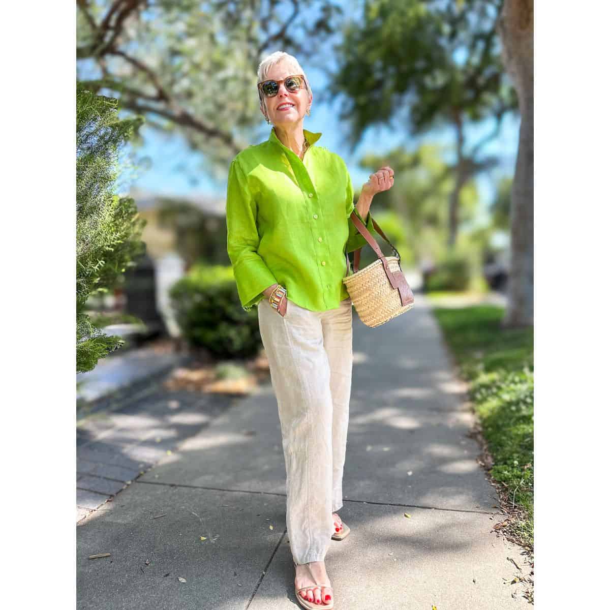 Susan B. wears a green linen shirt, undyed linen trousers, gold sandals, carries a basket bag.