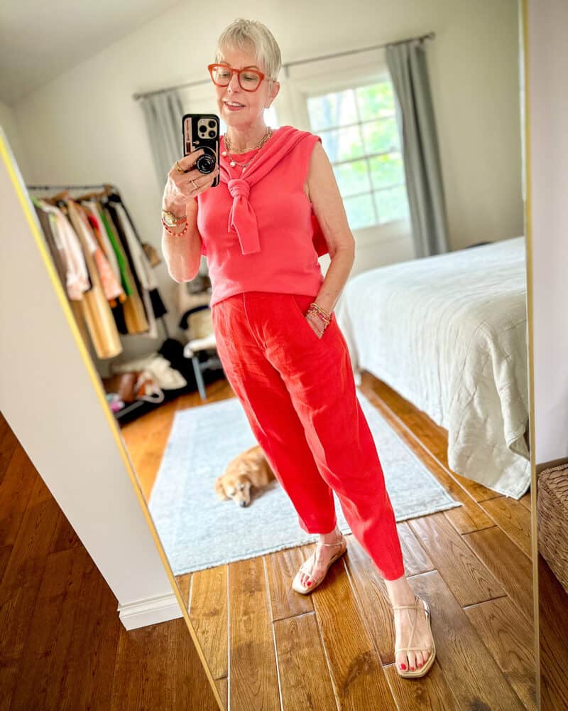 Susan B. wears a pink cashmere sleeveless sweater with matching cardigan tied over the shoulders, with red linen pants and gold strappy sandals.
